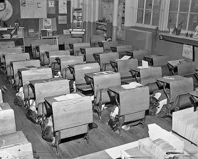 Original Caption: Open books are left on desks of the children of class 42-Junior High Elementary, as they dive under their desks during an "A" bomb drill.  Top view of children under their desks.